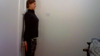 Photo of Emily Clarke standing with her back against the wall, demonstrating a posture exercise