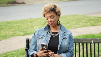 Image of a woman sat on a bench near grass, while reading from her phone