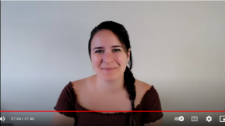 Zoe wears a brown top and smiles at the camera with a blank wall behind her