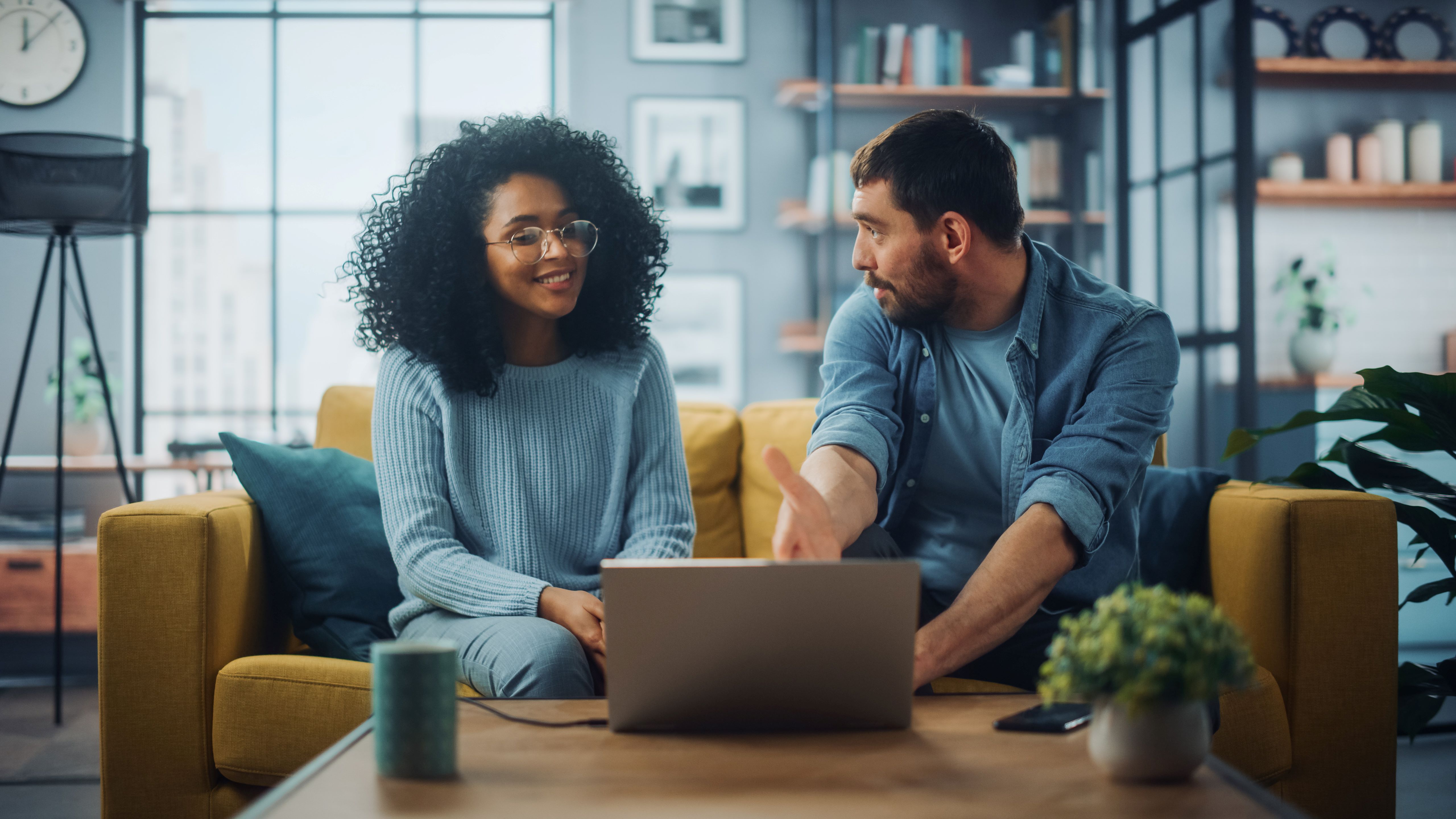 A man and a woman are sitting on a sofa, while using a laptop