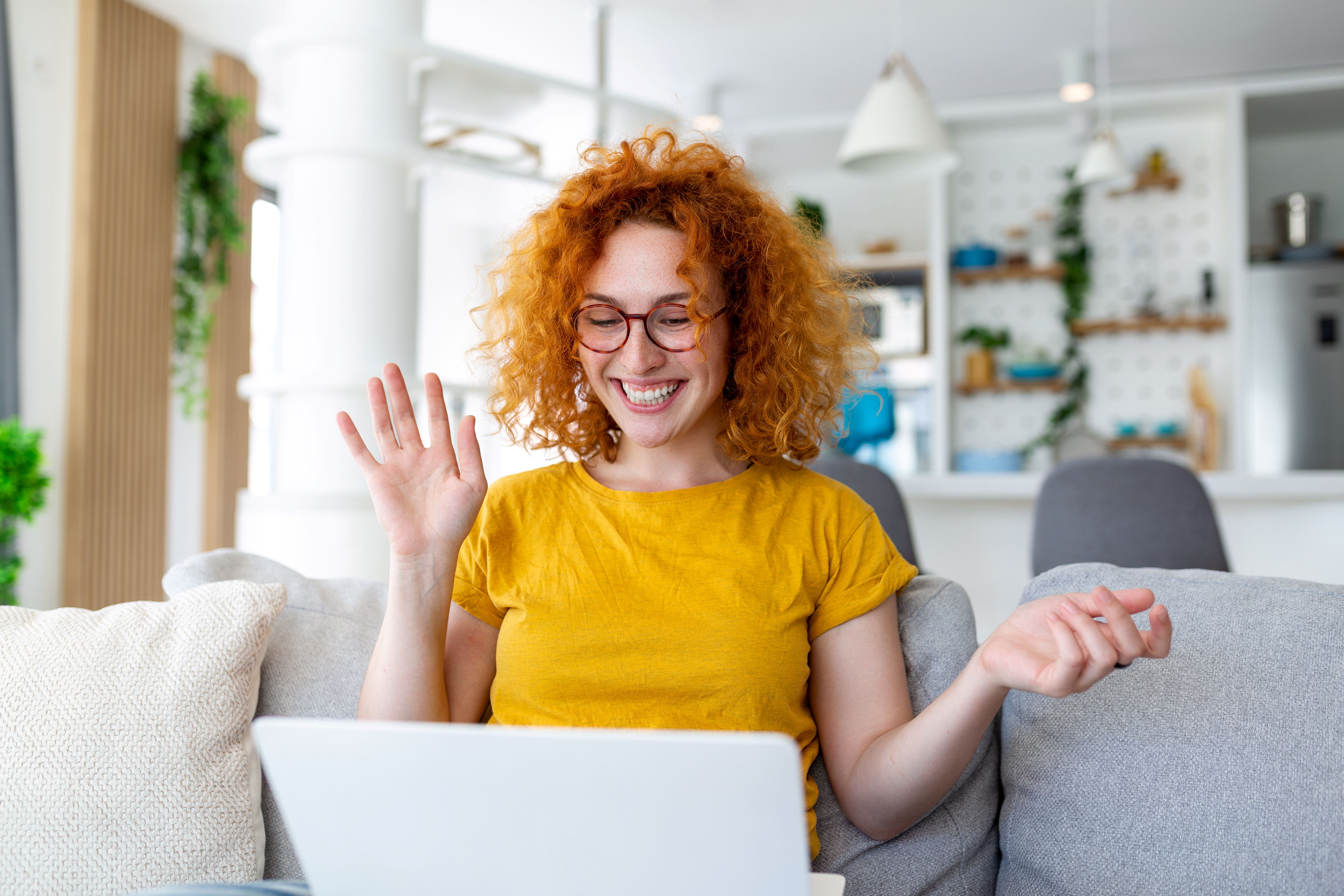 Woman with orange curly hair sits on a sofa, smiling and waving at a laptop screen